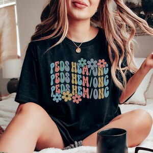 May include: Black t-shirt with a retro design featuring the text "PEDS HEM/ONG" repeated three times with colorful flowers in between each line.