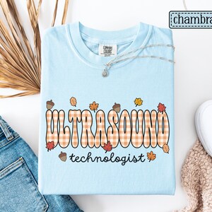 Puede incluir: Camiseta azul claro con las palabras "ULTRASOUND technologist" en una fuente retro con un patr&oacute;n de cuadros. El dise&ntilde;o incluye hojas de oto&ntilde;o y bellotas. Un collar plateado con un peque&ntilde;o colgante est&aacute; sobre la camiseta.