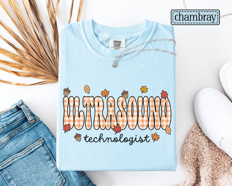 Puede incluir: Camiseta azul claro con las palabras "ULTRASOUND technologist" en una fuente retro con un patr&oacute;n de cuadros. El dise&ntilde;o incluye hojas de oto&ntilde;o y bellotas. Un collar plateado con un peque&ntilde;o colgante est&aacute; sobre la camiseta.