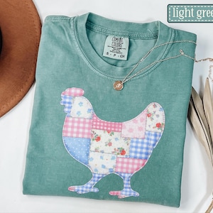 Peut inclure: T-shirt vert clair avec un motif de poulet en patchwork. Le poulet est fait de divers tissus à motifs, notamment des motifs floraux, vichy et de couleurs unies. Le t-shirt a un col rond et des manches courtes. Un collier en or avec un pendentif est sur le t-shirt.