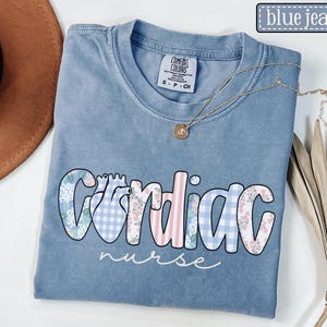 May include: A blue jean Comfort Colors t-shirt with the words "Cardiac nurse" in a floral and gingham design. A gold necklace with a pendant is draped across the shirt. The shirt is folded on a white surface.