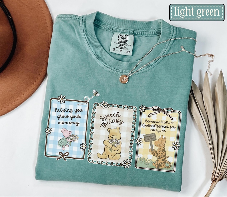 May include: Light green t-shirt with three framed graphics featuring Winnie the Pooh characters. The graphics include text that reads "helping you grow your own way," "Speech Therapy," and "Communication looks different for everyone."