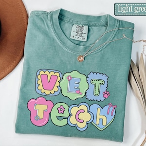 May include: Light green t-shirt featuring the words "VET TECH" in a colourful, bubble-style font. The design incorporates flowers, a paw print, and a heart. A silver necklace with a pendant is draped across the shirt.