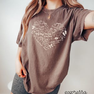 May include: Brown t-shirt with a heart-shaped design featuring the words "nicu nurse" and other text and illustrations. The shirt has short sleeves and a crew neck. The word "expressa" is at the bottom right.