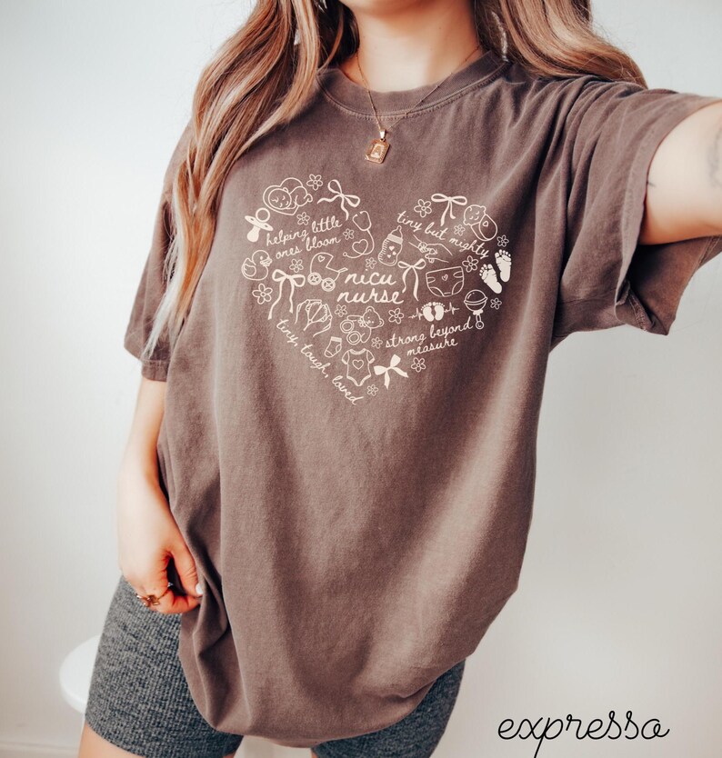 May include: Brown t-shirt with a heart-shaped design featuring the words "nicu nurse" and other text and illustrations. The shirt has short sleeves and a crew neck. The word "expressa" is at the bottom right.