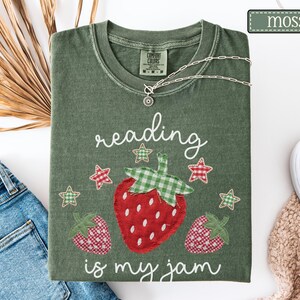 May include: A moss green t-shirt featuring a graphic of three strawberries with red and white gingham patterns and green gingham leaves. The text "reading is my jam" is printed in white script, accompanied by small red and green gingham stars.
