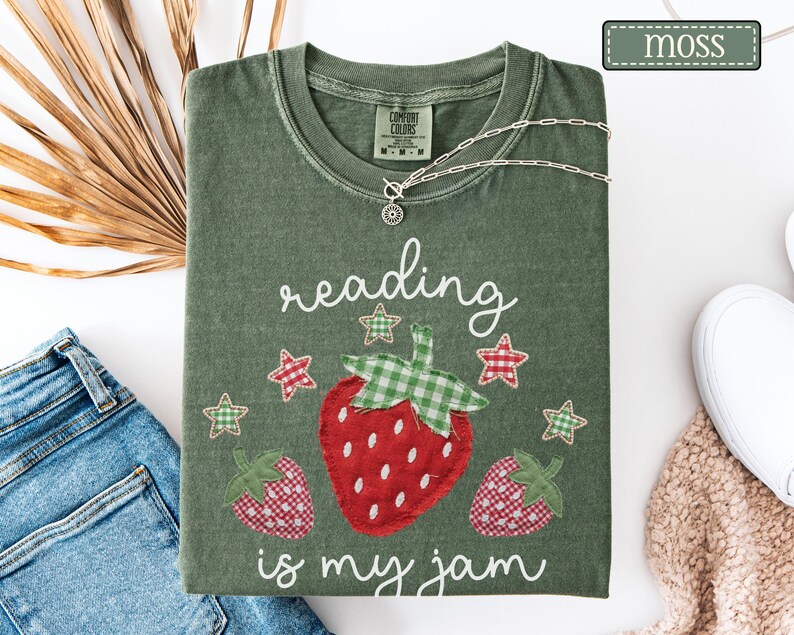 May include: A moss green t-shirt featuring a graphic of three strawberries with red and white gingham patterns and green gingham leaves. The text "reading is my jam" is printed in white script, accompanied by small red and green gingham stars.