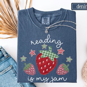 May include: A denim blue short-sleeve t-shirt featuring a graphic of three strawberries with green gingham leaves and red gingham bodies. The text reads "reading is my jam" in white script, surrounded by small red and green gingham stars.