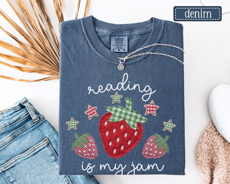 May include: A denim blue short-sleeve t-shirt featuring a graphic of three strawberries with green gingham leaves and red gingham bodies. The text reads "reading is my jam" in white script, surrounded by small red and green gingham stars.
