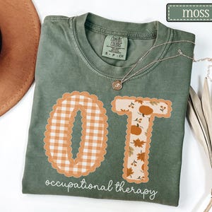 May include: A moss green t-shirt featuring the letters OT in a scalloped font. The O has an orange and white gingham pattern, and the T has a pumpkin and leaf print. The text occupational therapy is printed in white below the letters.
