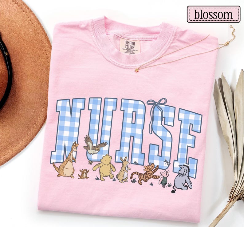 K&ouml;nnte beinhalten: Hellrosa T-Shirt mit dem Wort "NURSE" in blau-wei&szlig;em Vichy-Karo-Schriftzug. Unter dem Wort befinden sich Zeichentrickfiguren. Das Shirt ist von der Marke Comfort Colors.