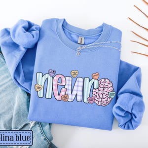 May include: Carolina blue sweatshirt with the word "neuro" in a colorful, playful font. The design includes a brain graphic and heart-shaped candies with phrases. A silver necklace rests on the shirt, which is paired with blue jeans.