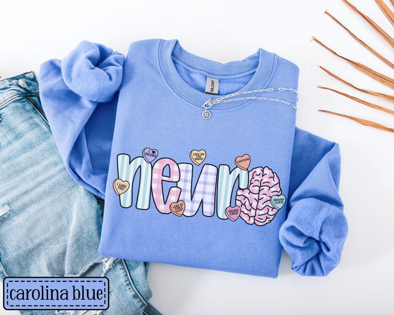 May include: Carolina blue sweatshirt with the word "neuro" in a colorful, playful font. The design includes a brain graphic and heart-shaped candies with phrases. A silver necklace rests on the shirt, which is paired with blue jeans.