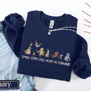 May include: Navy blue sweatshirt with a cartoon character graphic and the white text "SMALL STEPS STILL MOVE US FORWARD". The sweatshirt is displayed with blue jeans and a silver necklace.
