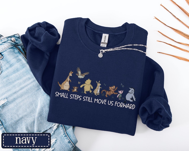 May include: Navy blue sweatshirt with a cartoon character graphic and the white text "SMALL STEPS STILL MOVE US FORWARD". The sweatshirt is displayed with blue jeans and a silver necklace.