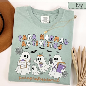May include: A sage green t-shirt featuring the text "PARA NORMAL ACTIVITIES" in purple and orange, with a graphic of three ghosts holding a clipboard, a coffee cup, and a walkie-talkie, and the text "paraprofessional" in orange at the bottom.