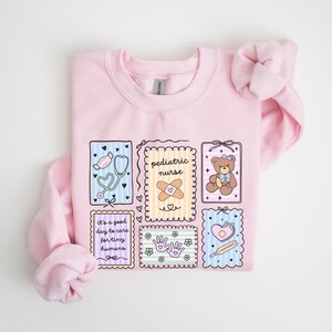 May include: A light pink sweatshirt with a colorful graphic design featuring medical-themed illustrations. The design includes a stethoscope, band-aid, teddy bear, and the words "pediatric nurse" and "It's a good day to care for tiny humans."