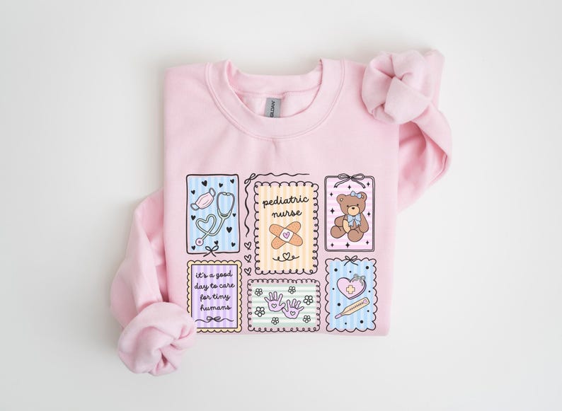 May include: A light pink sweatshirt with a colorful graphic design featuring medical-themed illustrations. The design includes a stethoscope, band-aid, teddy bear, and the words "pediatric nurse" and "It's a good day to care for tiny humans."