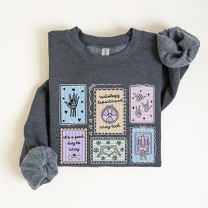 May include: A dark gray sweatshirt featuring several colorful, framed illustrations related to radiology. The illustrations include a hand, a peace sign, a skeleton, and the text "radiology department x-ray tech" and "it's a good day to xray".