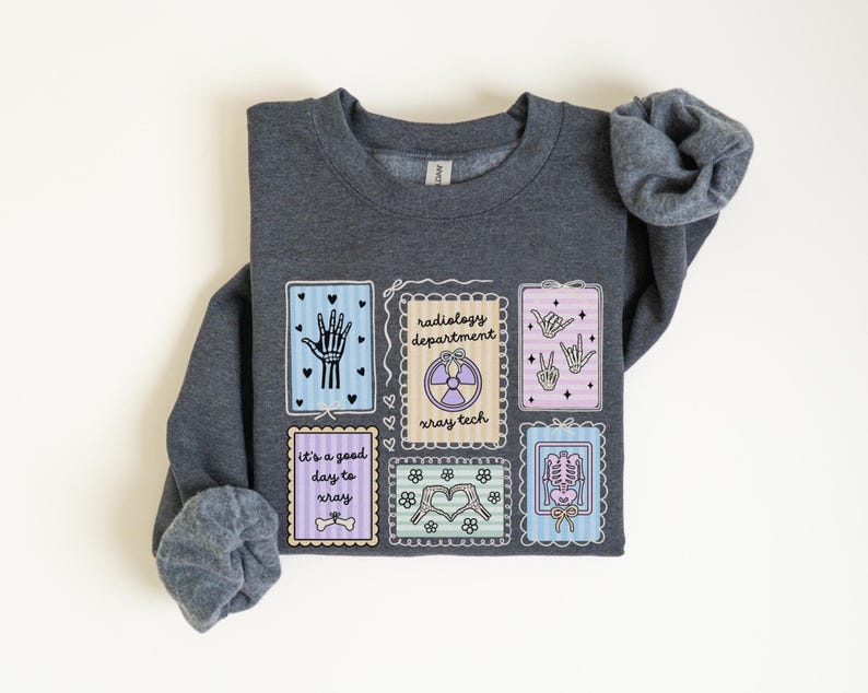May include: A dark gray sweatshirt featuring several colorful, framed illustrations related to radiology. The illustrations include a hand, a peace sign, a skeleton, and the text "radiology department x-ray tech" and "it's a good day to xray".