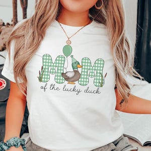 May include: White t-shirt with a green and white checkered design that says "Mim of the lucky duck" with a cartoon duck wearing a party hat and holding a balloon.
