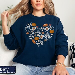 May include: A navy blue crewneck sweatshirt featuring a heart-shaped graphic composed of various icons including pumpkins, flowers, radiation symbols, a skeleton hand, a medical caduceus, and an X-ray, with the word Radiology printed in the center.