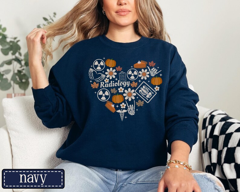 May include: A navy blue crewneck sweatshirt featuring a heart-shaped graphic composed of various icons including pumpkins, flowers, radiation symbols, a skeleton hand, a medical caduceus, and an X-ray, with the word Radiology printed in the center.