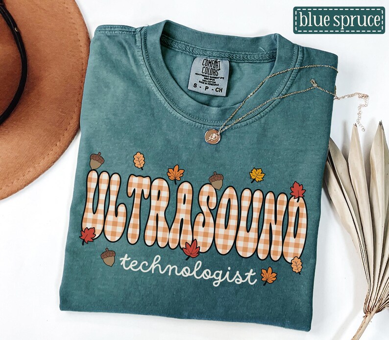 Puede incluir: Una camiseta Comfort Colors de color verde azulado con las palabras "ULTRASOUND" en un dise&ntilde;o a cuadros y "technologist" en escritura blanca. La camiseta presenta gr&aacute;ficos con tem&aacute;tica oto&ntilde;al, que incluyen bellotas y hojas. Un collar dorado est&aacute; drapeado sobre la camiseta.