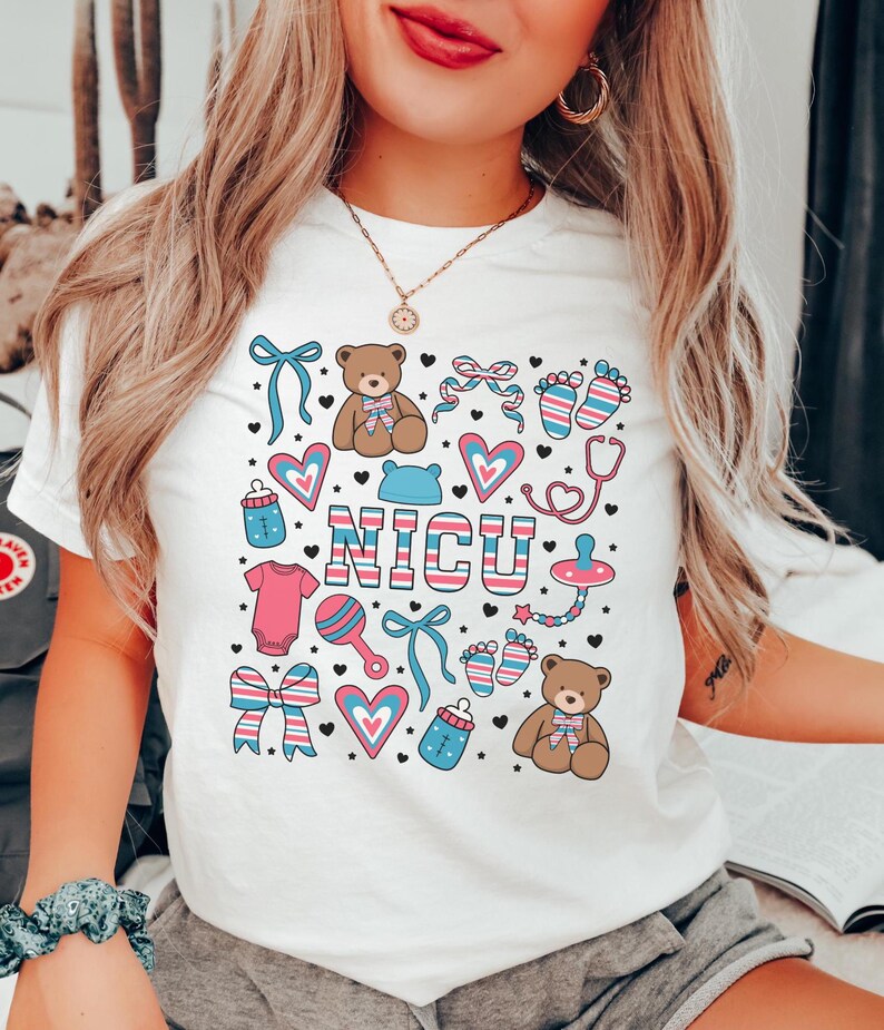 May include: White t-shirt with a pink, blue, and white graphic design featuring a teddy bear, baby bottle, rattle, and the word "NICU".