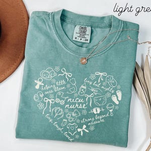 May include: A light green t-shirt with a heart-shaped design featuring baby-related illustrations and the words "nicu nurse." The shirt also has the phrases "helping little ones bloom," "tiny but mighty," and "strong beyond measure." A silver necklace is draped across the shirt.