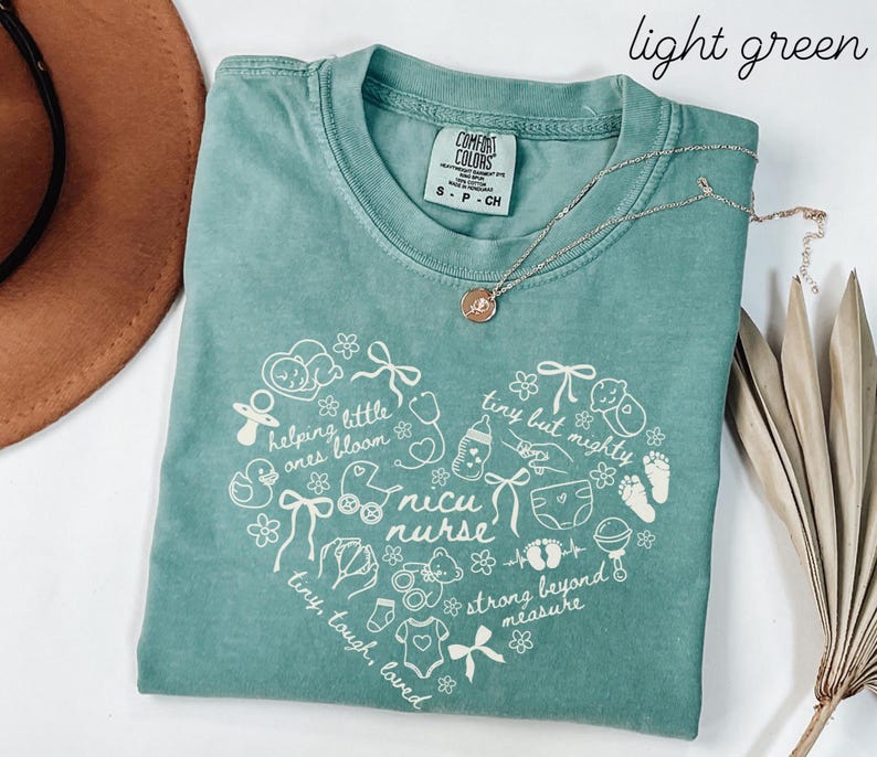May include: A light green t-shirt with a heart-shaped design featuring baby-related illustrations and the words "nicu nurse." The shirt also has the phrases "helping little ones bloom," "tiny but mighty," and "strong beyond measure." A silver necklace is draped across the shirt.
