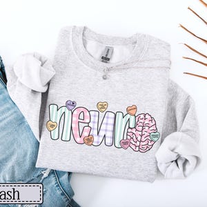 May include: A light gray sweatshirt with the word "NEURO" in a colorful, playful font, and a brain graphic. The letters are decorated with heart-shaped candies. The word "ash" is printed on a label.