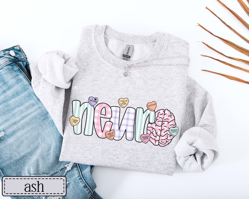 May include: A light gray sweatshirt with the word "NEURO" in a colorful, playful font, and a brain graphic. The letters are decorated with heart-shaped candies. The word "ash" is printed on a label.