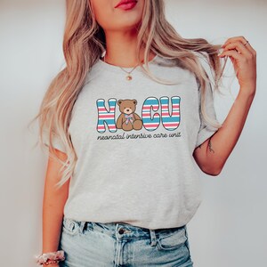 Puede incluir: Una camiseta gris con un gr&aacute;fico de un osito de peluche con un lazo rosa y el texto "NICU" en rayas azules, rosas y blancas. El texto "unidad de cuidados intensivos neonatales" est&aacute; escrito debajo del gr&aacute;fico.