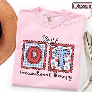 May include: A light pink t-shirt with the words "OT Occupational Therapy" printed on the front. The letters "OT" are inside of red postage stamp designs. The "O" is red with a dotted design, and the "T" is blue with a floral pattern.