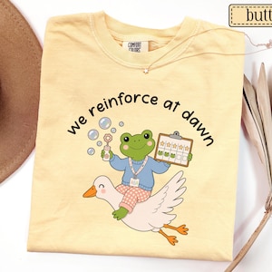 May include: A butter yellow t-shirt featuring a graphic of a green frog wearing a blue sweater and pink checkered pants, riding a white goose. The frog holds a bubble wand and a clipboard with star stickers. The text reads, we reinforce at dawn.