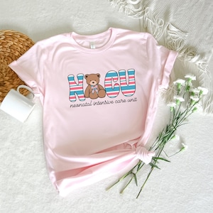Puede incluir: Camiseta rosa con un gr&aacute;fico de un osito de peluche y el texto "NICU" con rayas azules y rosas. El texto debajo del gr&aacute;fico dice "unidad de cuidados intensivos neonatales".