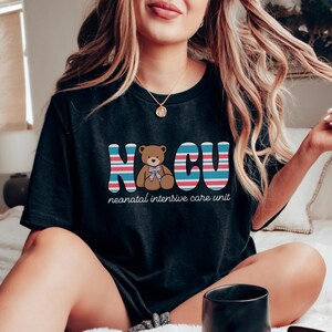 Puede incluir: Una camiseta negra con un gr&aacute;fico de un osito de peluche con un lazo azul y el texto "NICU" en letras de colores. El texto debajo del gr&aacute;fico dice "unidad de cuidados intensivos neonatales".