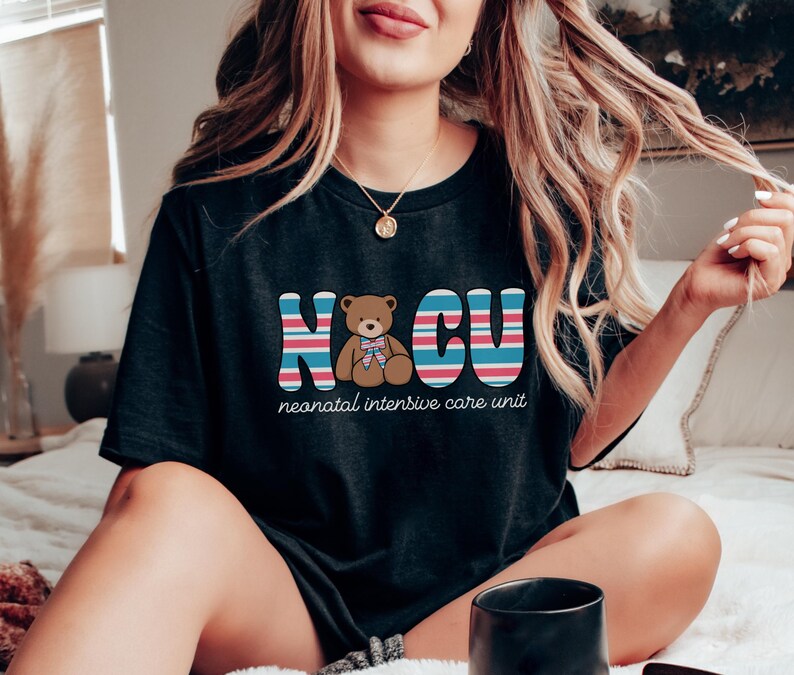 Puede incluir: Una camiseta negra con un gr&aacute;fico de un osito de peluche con un lazo azul y el texto "NICU" en letras de colores. El texto debajo del gr&aacute;fico dice "unidad de cuidados intensivos neonatales".