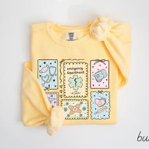 Pode incluir: Camisola amarela clara com um gr&aacute;fico com s&iacute;mbolos m&eacute;dicos e as palavras "emergency department". O design inclui um estetosc&oacute;pio, uma m&aacute;scara facial e outras ilustra&ccedil;&otilde;es relacionadas com a medicina. A palavra "butter" est&aacute; escrita em it&aacute;lico no canto inferior direito.