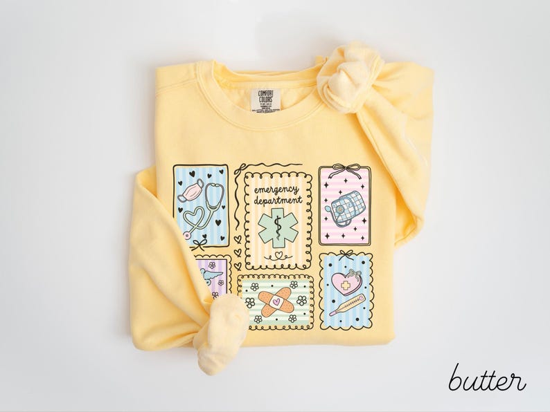 Pode incluir: Camisola amarela clara com um gr&aacute;fico com s&iacute;mbolos m&eacute;dicos e as palavras "emergency department". O design inclui um estetosc&oacute;pio, uma m&aacute;scara facial e outras ilustra&ccedil;&otilde;es relacionadas com a medicina. A palavra "butter" est&aacute; escrita em it&aacute;lico no canto inferior direito.