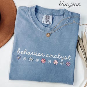 May include: A blue jean colored t-shirt with the words "behavior analyst" in white script, decorated with small flower graphics in blue, pink, and purple. A gold necklace with a pendant rests on the shirt.