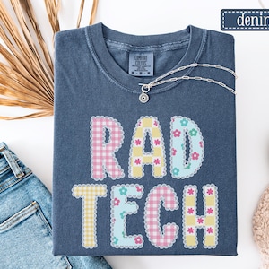 May include: A denim blue t-shirt with the words "RAD TECH" in a patchwork design. The letters are decorated with gingham and floral patterns in pastel colors. A silver necklace with a pendant is on the shirt.
