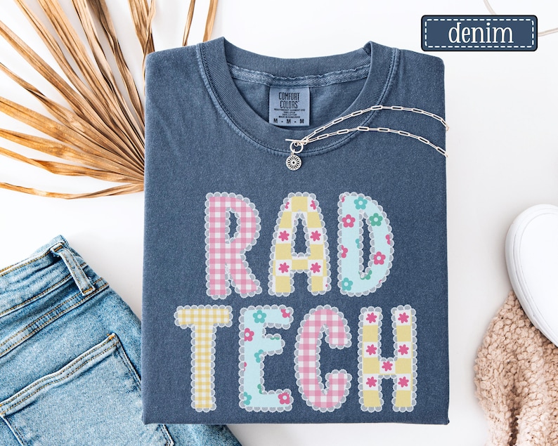 May include: A denim blue t-shirt with the words "RAD TECH" in a patchwork design. The letters are decorated with gingham and floral patterns in pastel colors. A silver necklace with a pendant is on the shirt.