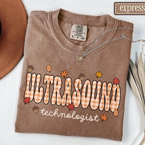 Puede incluir: Camiseta marr&oacute;n con la frase "ULTRASOUND technologist" en una fuente retro con un dise&ntilde;o de cuadros, decorada con hojas de oto&ntilde;o y bellotas. Se ve un collar plateado con un colgante en la camiseta.