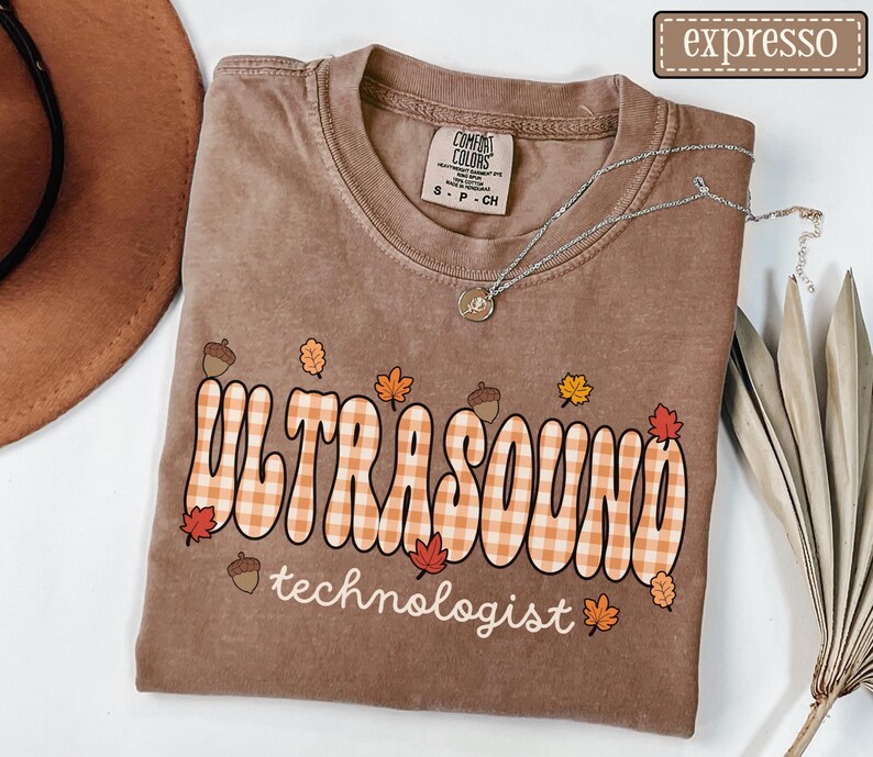 Puede incluir: Camiseta marr&oacute;n con la frase "ULTRASOUND technologist" en una fuente retro con un dise&ntilde;o de cuadros, decorada con hojas de oto&ntilde;o y bellotas. Se ve un collar plateado con un colgante en la camiseta.