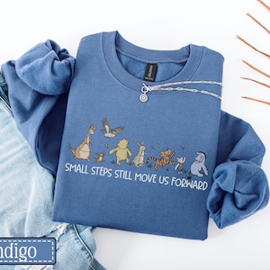 May include: Indigo blue crew neck sweatshirt with a cartoon illustration of characters including a kangaroo, bear, tiger, and pig. The text "SMALL STEPS STILL MOVE US FORWARD" is printed below the characters. A silver necklace is draped across the sweatshirt.