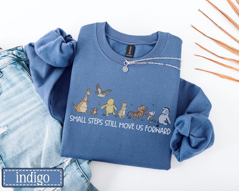 May include: Indigo blue crew neck sweatshirt with a cartoon illustration of characters including a kangaroo, bear, tiger, and pig. The text "SMALL STEPS STILL MOVE US FORWARD" is printed below the characters. A silver necklace is draped across the sweatshirt.