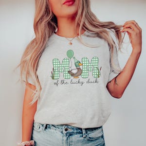 May include: A light gray t-shirt with a green and white checkered design that says "Mom of the lucky duck" with a cartoon duck wearing a party hat and holding a balloon.
