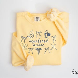 May include: A light yellow sweatshirt with the words "registered nurse" surrounded by medical-themed illustrations in navy blue. The sweatshirt is knotted at the bottom and has a matching bow on the shoulder.